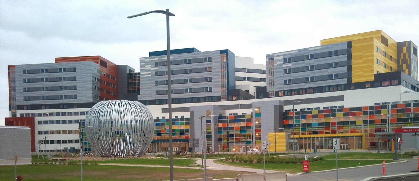 The McGill University Health Centre superhospital in Montreal: colourful multi-storey hospital blocks behind a large open-lattice metal sculpture.