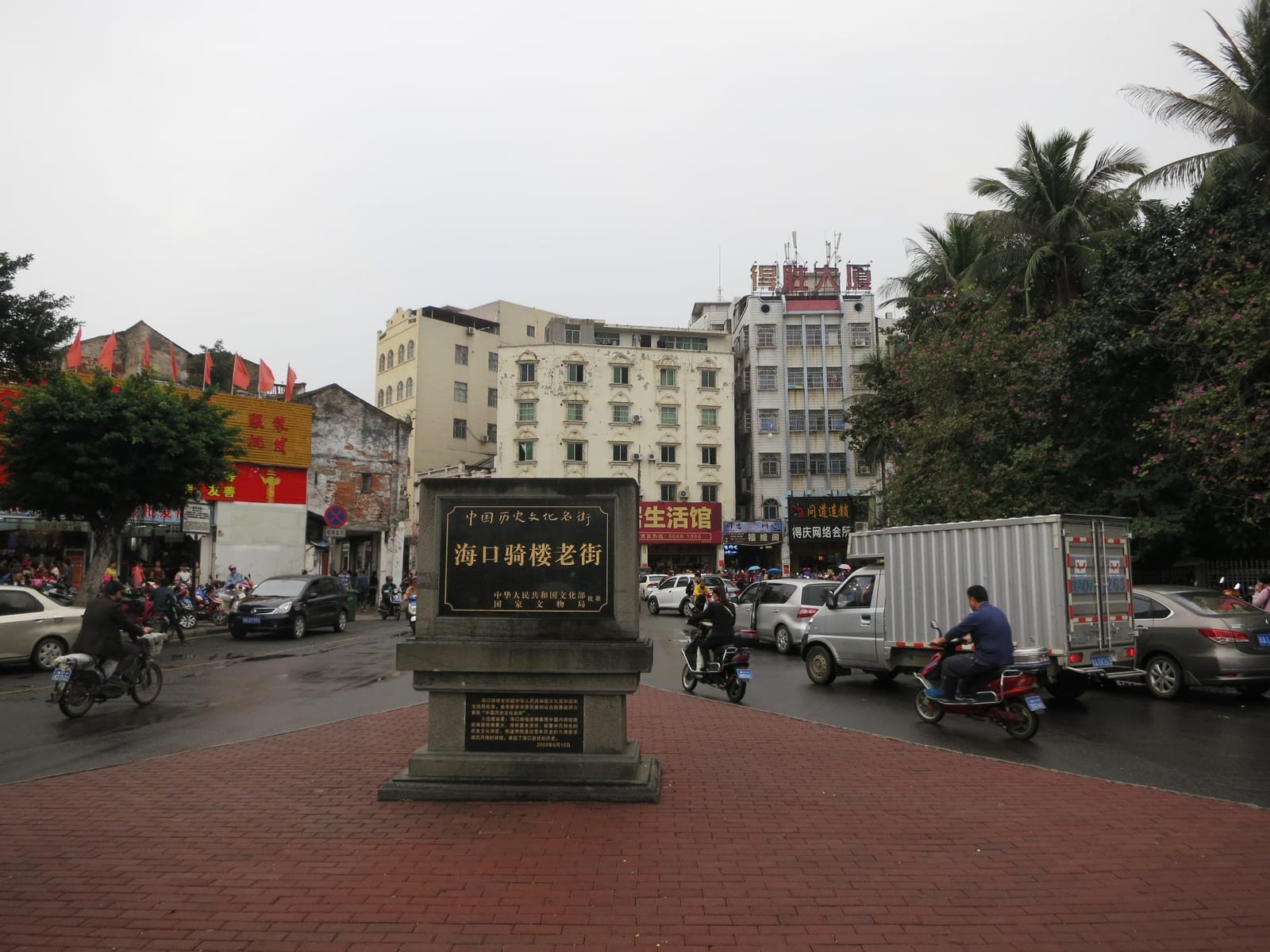 Illustrative image: street scene in Haikou, Hainan, with a stone marker, scooters and shophouses.